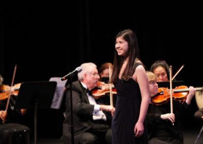 A girl in a black dress singing in front of the SoCal Philharmonic orchestra.