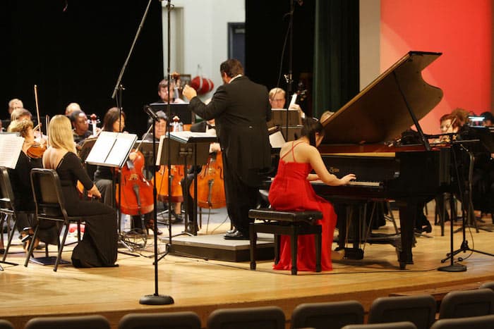 A woman in a red dress is playing a piano in front of an orchestra.